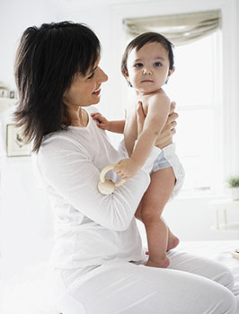 Baby standing on mother's lap. Mother is hoding baby's hands and smiling.