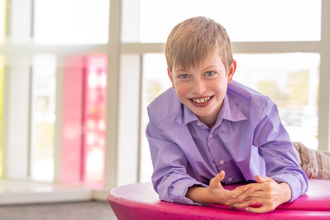 Smiling blonde kid with green eyes and wearing a purple shirt.