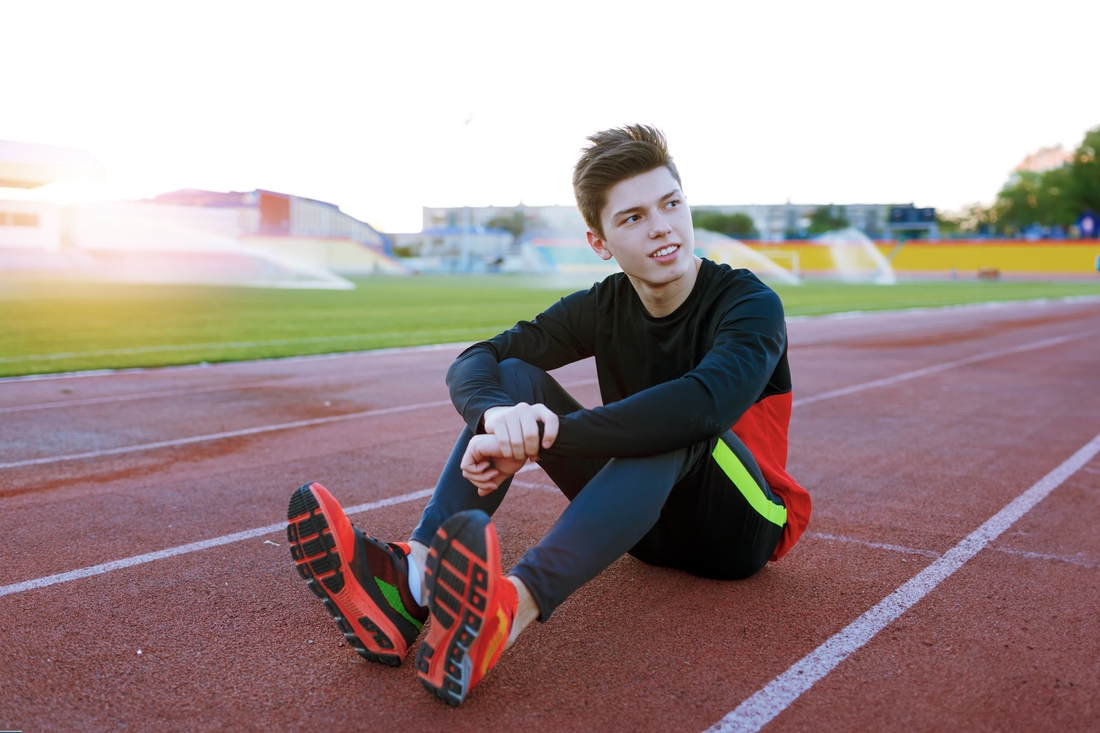 Older teen boy sitting on a track wearing cleats looking up.