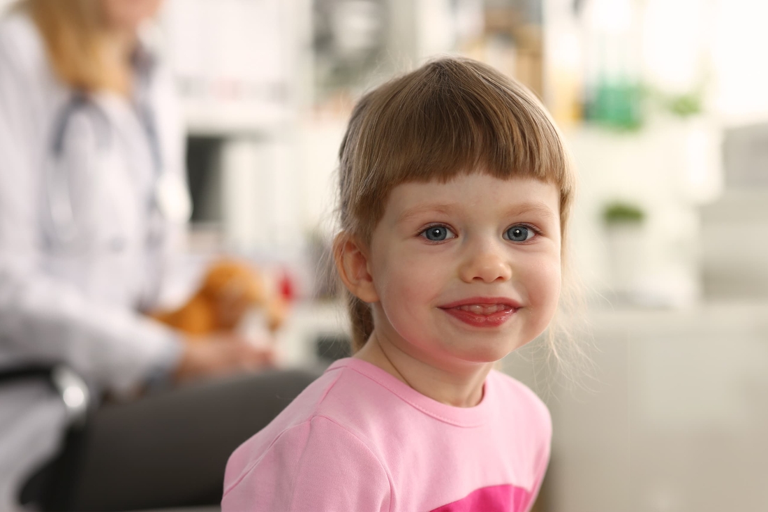 Girl with bangs smiling with doctor blurred in background.