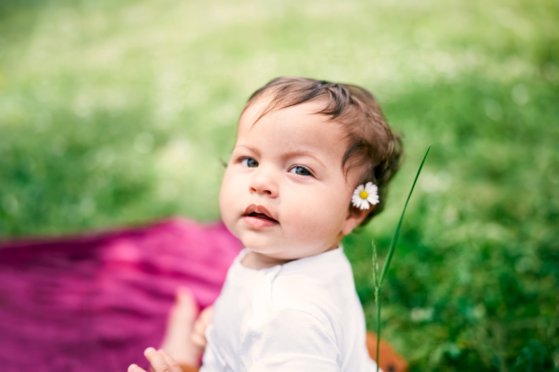 Infant girl sitting on pink blanket with flower tucked behind her ear at a park.