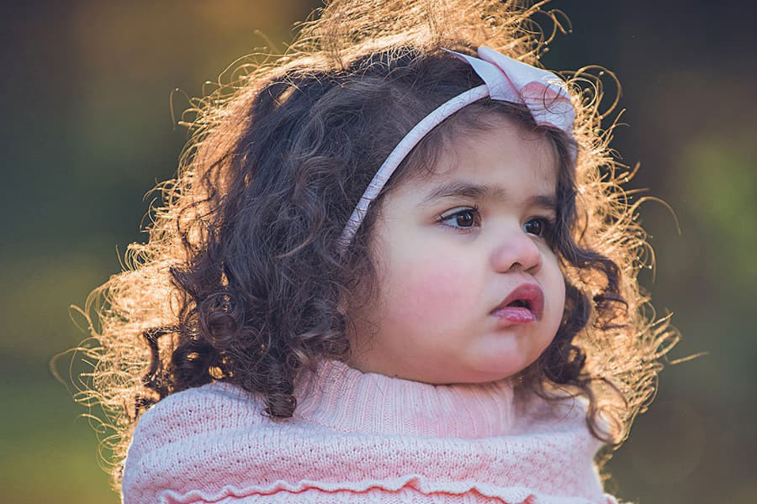 Little girl with brown curly hair and pink bow looks to the side.