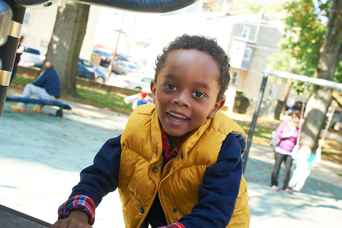 Little boy in yellow vest on the playground smiling at the camera.