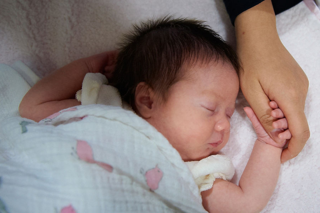 Baby with black hair sleeping under blanket holding adult's hand.