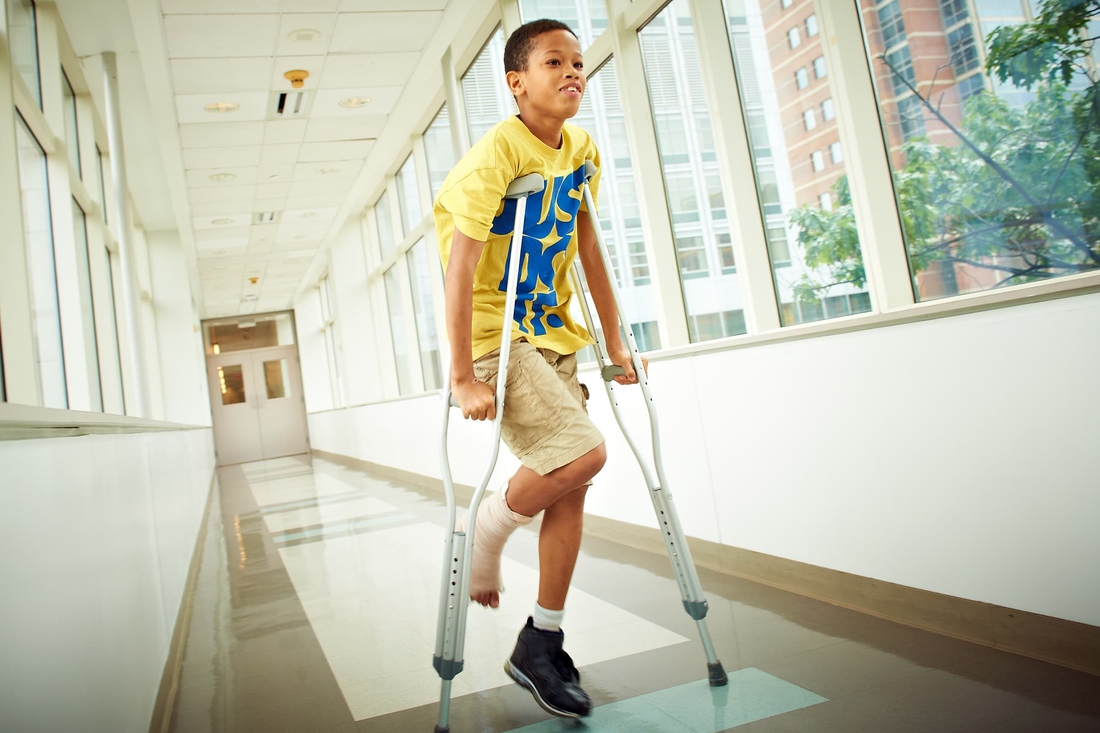 School aged boy with a bandaged foot/leg using crutches to walk down a sunny hallway
