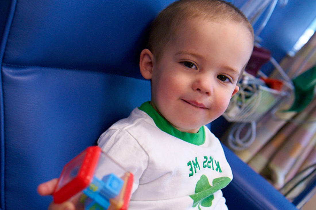 Little boy sits on blue hospital chair and looks at camera.