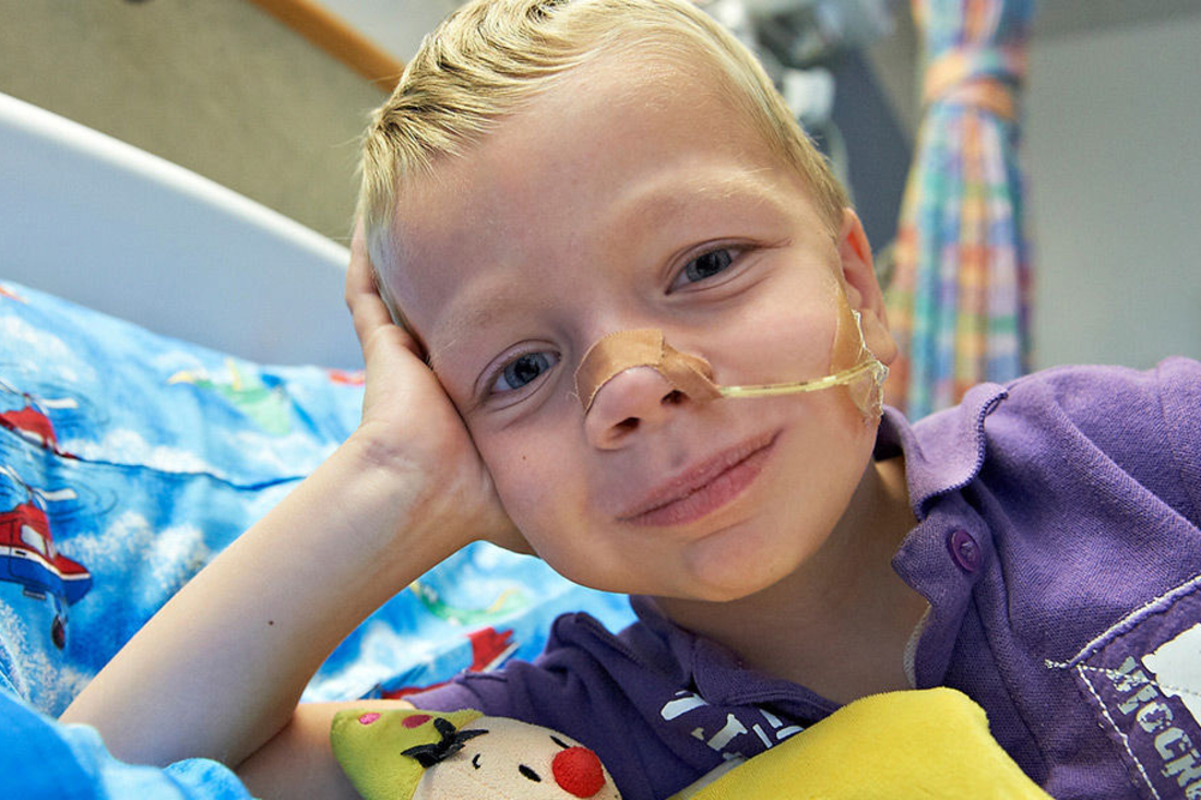 Young blonde boy with tube over his nose and cheek rests on his hand in a hospital bed.