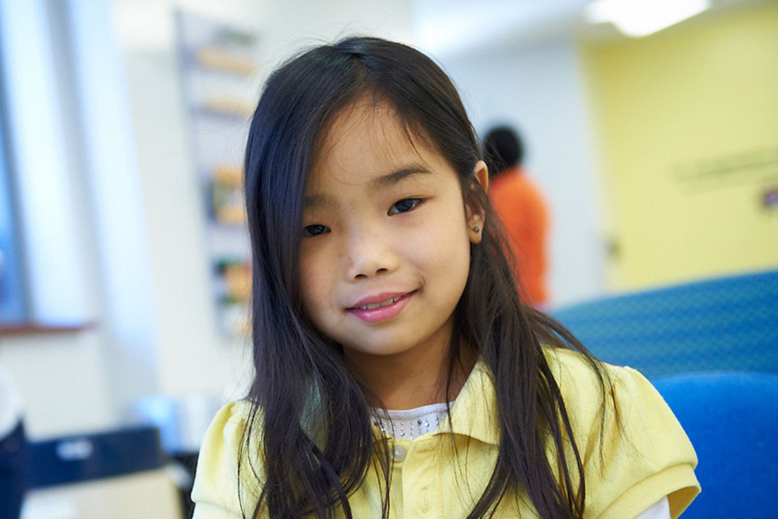 Young Asian girl with long dark hair and wearing a yellow polo smiles at camera.