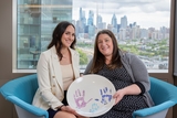 Two women sitting holding up a ceramic memory plate with handprints made by a family where the child is now deceased