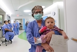 Nurse carrying infant walking in hospital corridor with other nurses in background