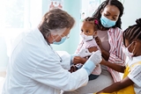 Physician examining a child in a mask who is sitting on mom's lap.