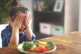 Young girl with hands over eyes sitting in front of a plate of food
