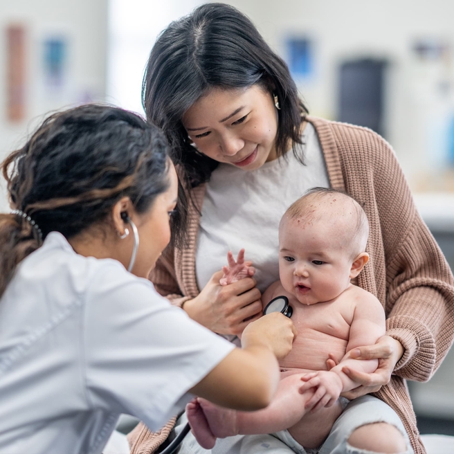 Doctor listening to heart and giving exam to baby sitting on her mothers lap.
