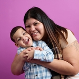 mom smiling and hugging her boy with a purple background behing them.