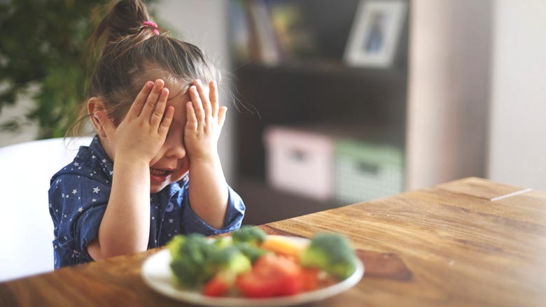 Young girl with hands over eyes sitting in front of a plate of food